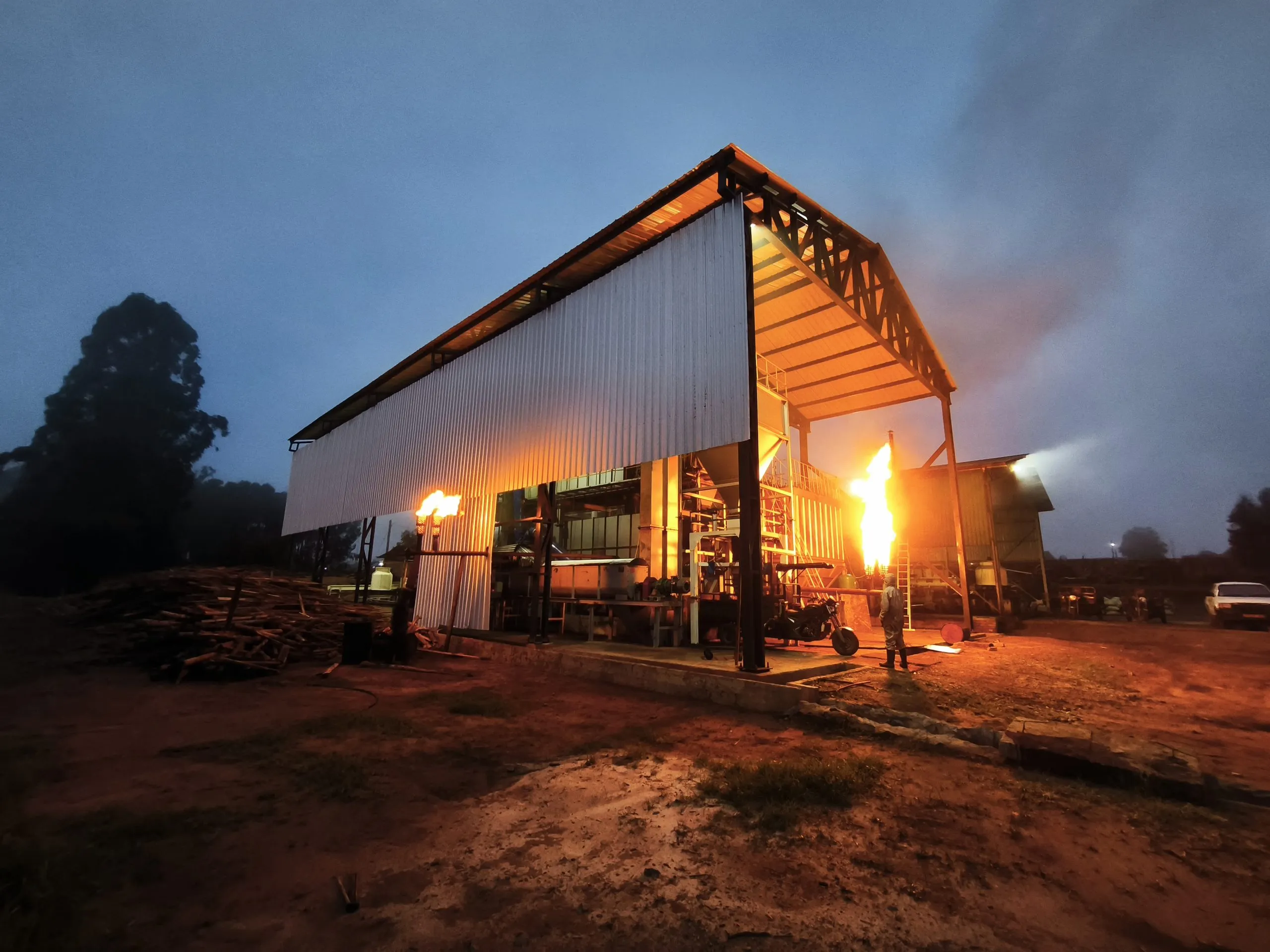 A pyrolysis plant running at dusk, its flare alight, with stacked feedstock alongside